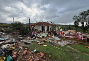 Casa destruída por tempestade em área rural, com telhado danificado, árvores derrubadas e destroços espalhados pelo terreno.